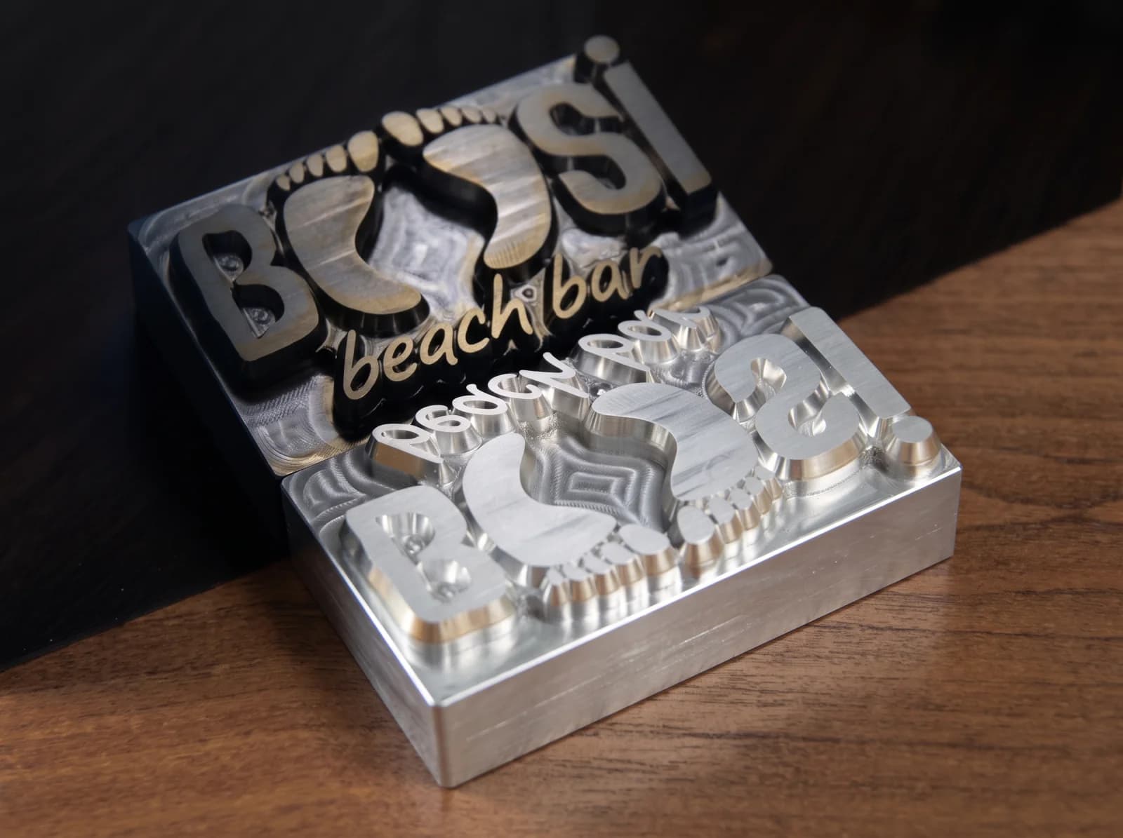 A freshly CNC-milled aluminium branding stamp for Bossi Beach Bar, the machine's toolpaths still visible on the face, reflected in a dark surface